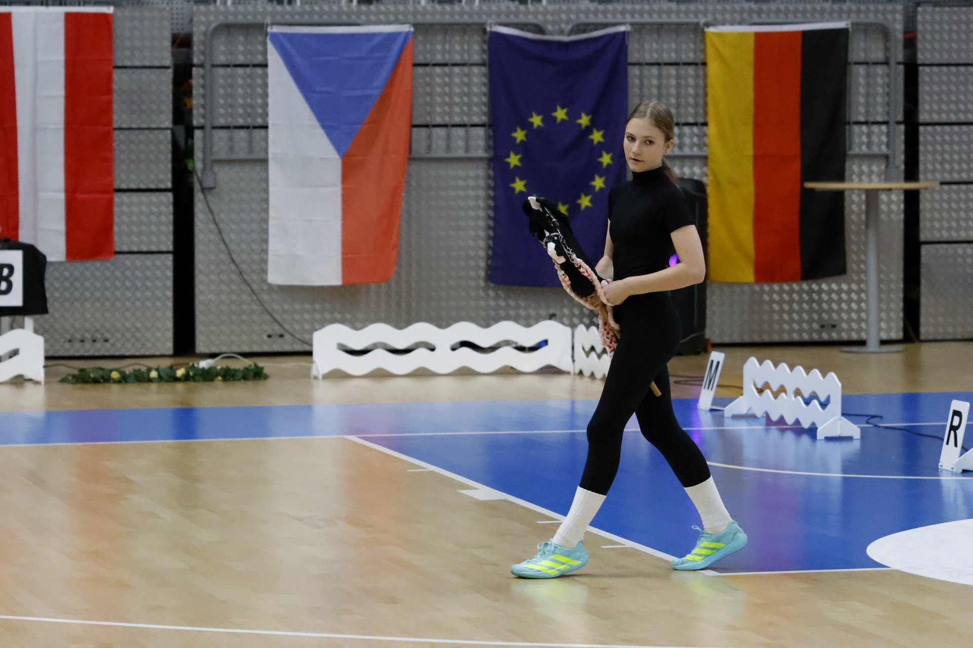 Závodnice v černém předvádí drezurní úlohu s Hobby Horse koněm v drezurním obdélníku před vlajkami evropských zemí.